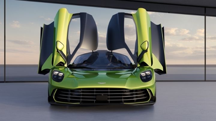 Green Aston Martin Valhalla with both front doors open parked in front of a glass wall with the sky in the background