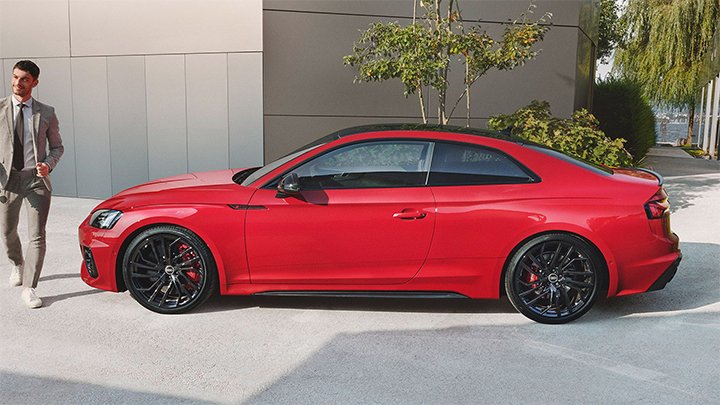 Side of red Audi RS5 Coupé parked in front of a building with grey walls and a man wearing a grey suit is walking in front of it