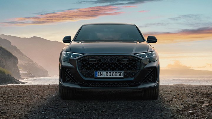 Front of Audi RSQ8 parked on the sand with an ocean and mountain in the background during a sunset
