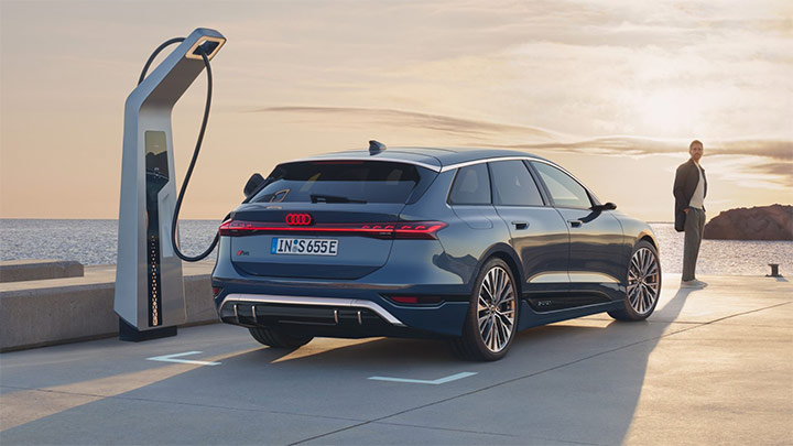 Back of a blue Audi S6 Avant e-Tron parked in the asphalt with the ocean and mountain in the background whilst plugged into the charger and with a man standing in front of it
