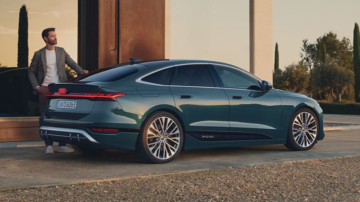 Man wearing a white shirt and grey blazer leaning on the blue S6 Sportback e-Tron that is parked in front of a modern house with large mirrored glass walls and trees on the right