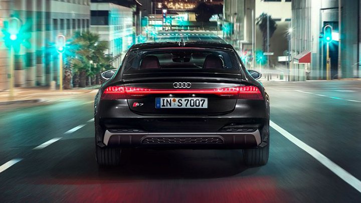 Back of a black Audi S7 Sportback driving in a large city with several buildings and green lights ahead