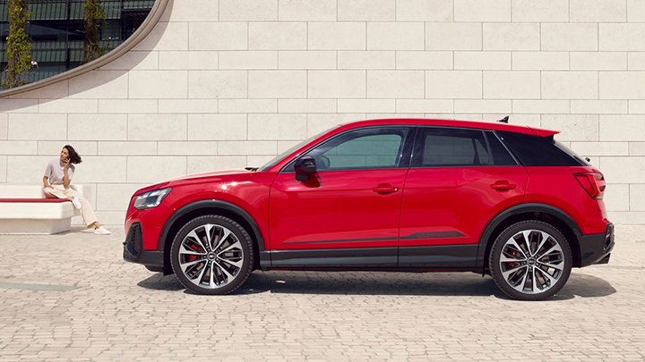 The side of a red Audi SQ2 parked in front of a beige wall with a woman wearing beige trousers and grey shirt sitting on a bench in front of the car and the wall