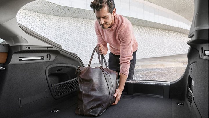 View from Audi SQ5 boot with a man wearing a pink sweater and black trousers placing a dark brown leather duffle bag in it. The car is parked in front of a wall made with white tiles