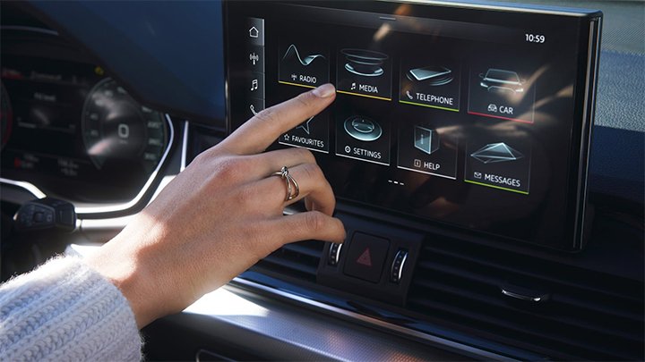Image of Audi SQ5 infotainment system with the menu icons displayed on the scree. A hand of a woman wearing white sweater and a gold ring is reaching to select an option from the menu