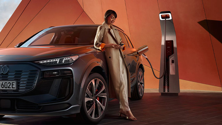 A man wearing a long beige trench coat leaning on a grey Audi SQ6 e-Tron whilst the car is plugged into the charger. An orange modern building can be seen in the background