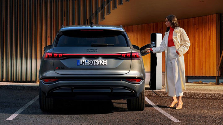 Woman wearing white trousers, orange top and beige trench coat plugging her Audi SQ6 e-Tron onto the charger. A modern house with details in varnish wood can be seen in the background.