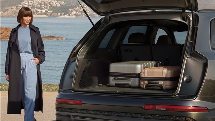 Audi SQ7 Sportback boot with a small and a large suitcase in it. A woman is standing next to it and the car is parked in front of the ocean