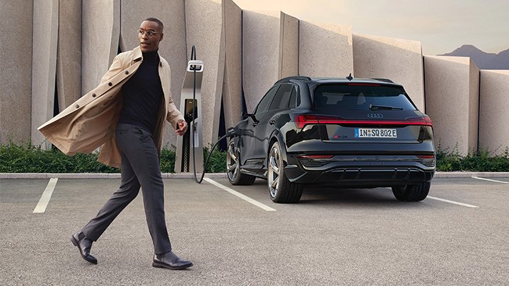 Man wearing black shirt, grey trousers and beige trench coat walking in front of a black Audi SQ8 e-tron that is parked with its back to the camera in front of a beige wall whilst plugged in to the charger
