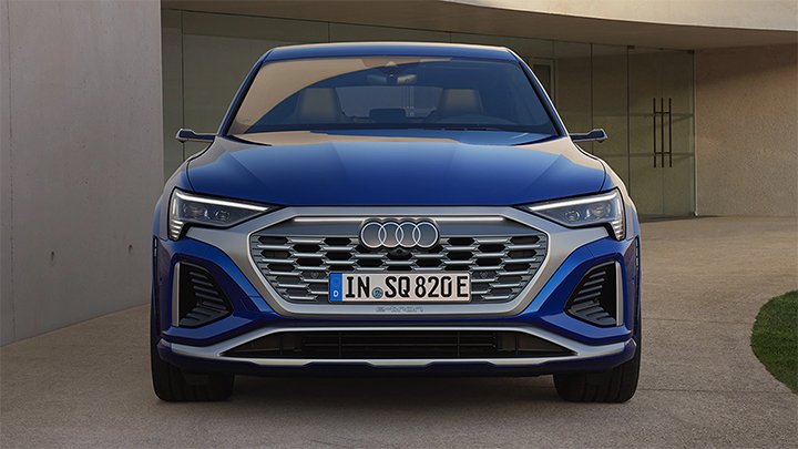 Front of blue Audi SQ8 Sportback e-Tron displaying the vehicle's grille and Audi's iconic logo. Parked in front of a beige garage.