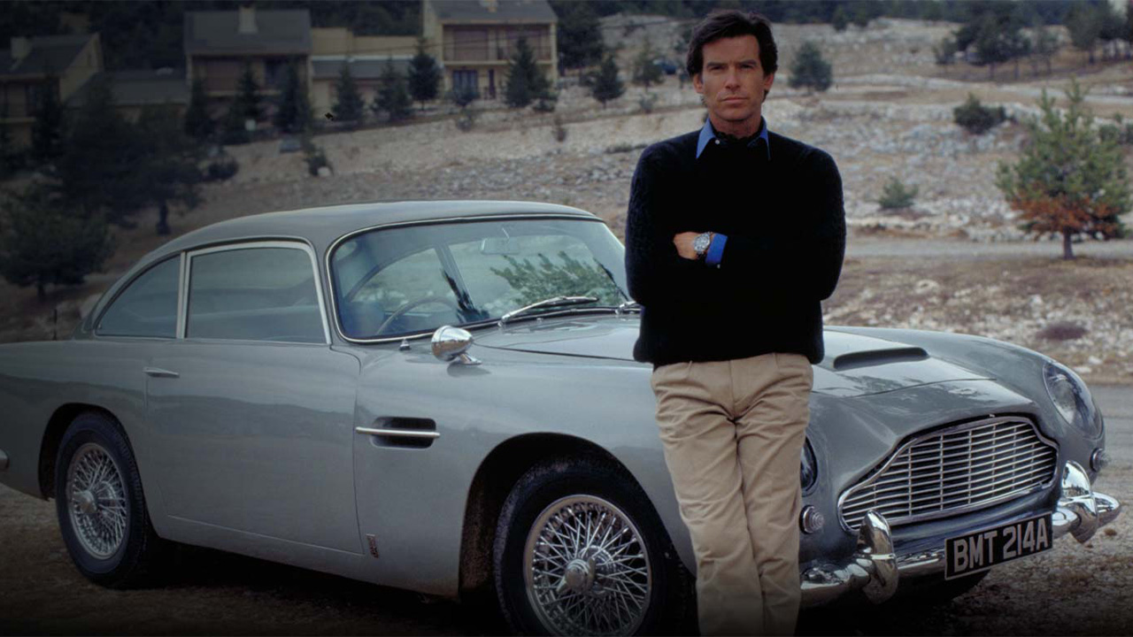 Pierce Brosnan leaning against an Aston Martin DB5
