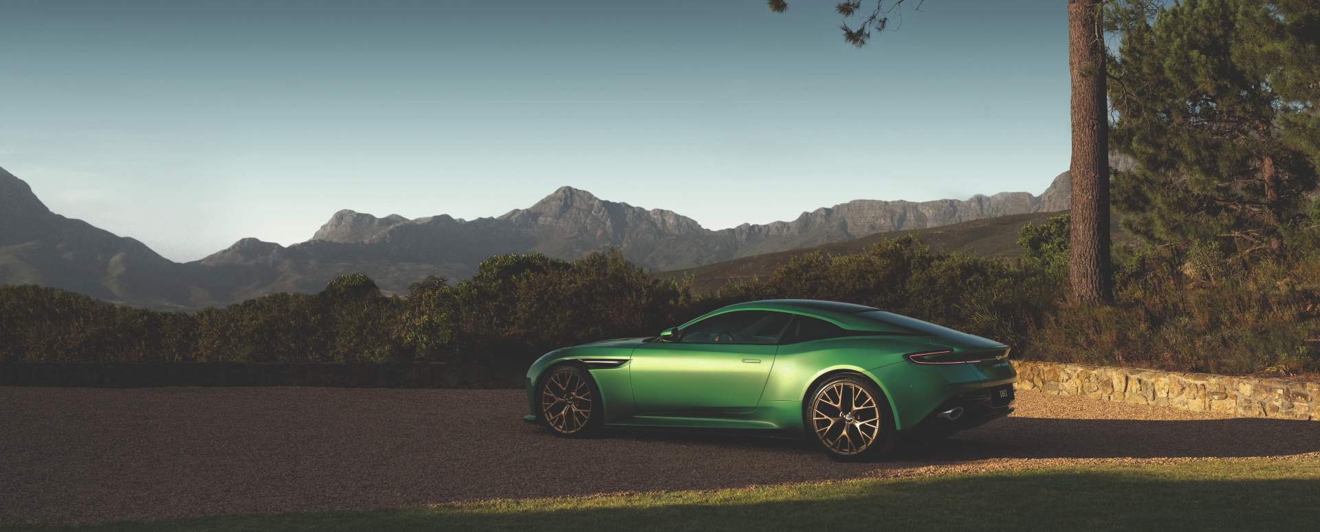 Green Aston Martin DB12 grand tourer shown in side profile against a mountainous landscape