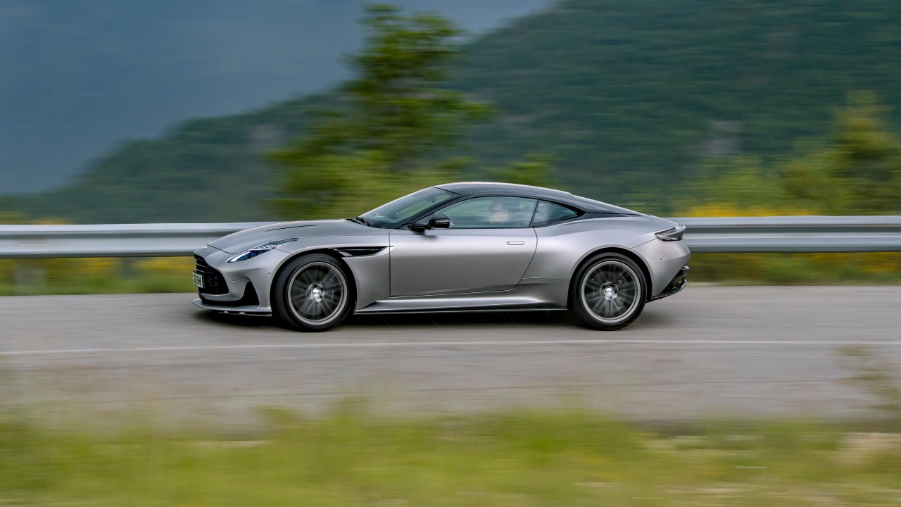 Aston Martin DB12 grand tourer driving from a side angle on a rural road