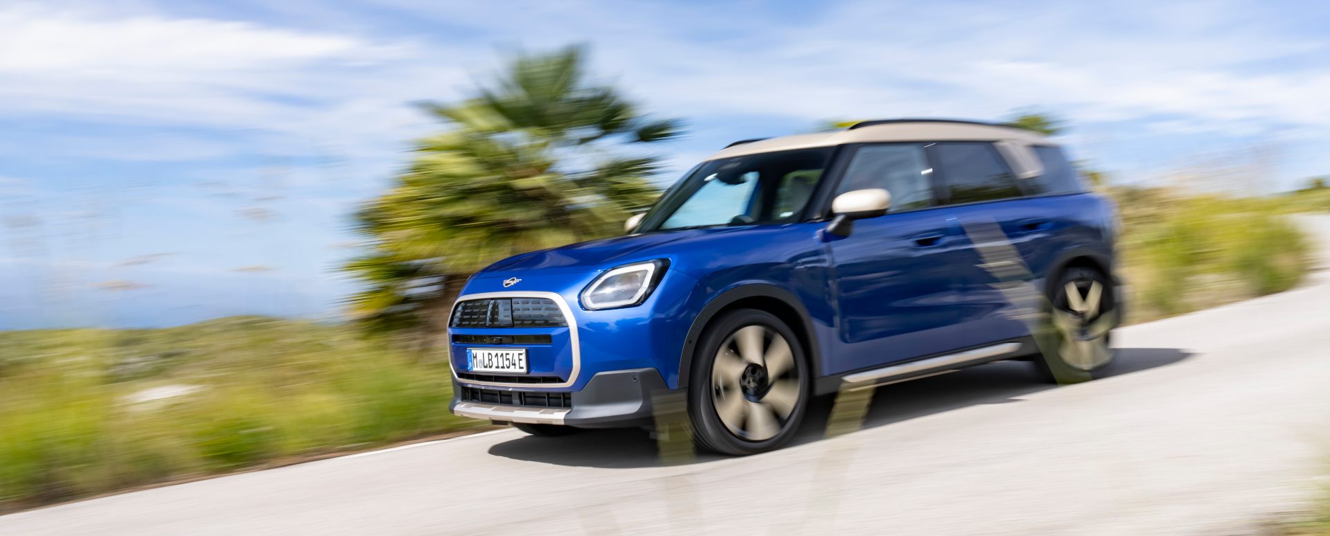 Blue MINI Countryman electric SUV driving from a front three‑quarter angle on a country road