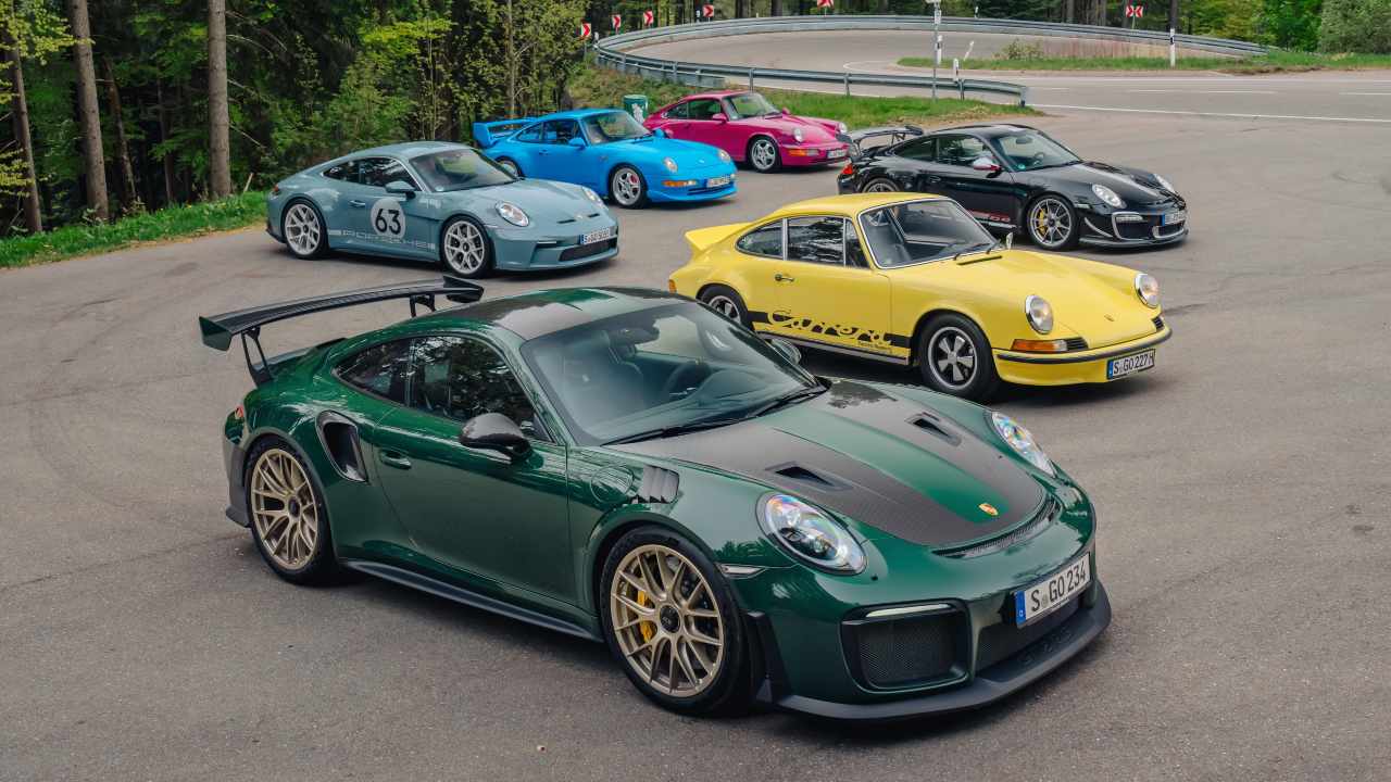 Multiple generations of Porsche 911 sports cars in various colours parked together on a country road