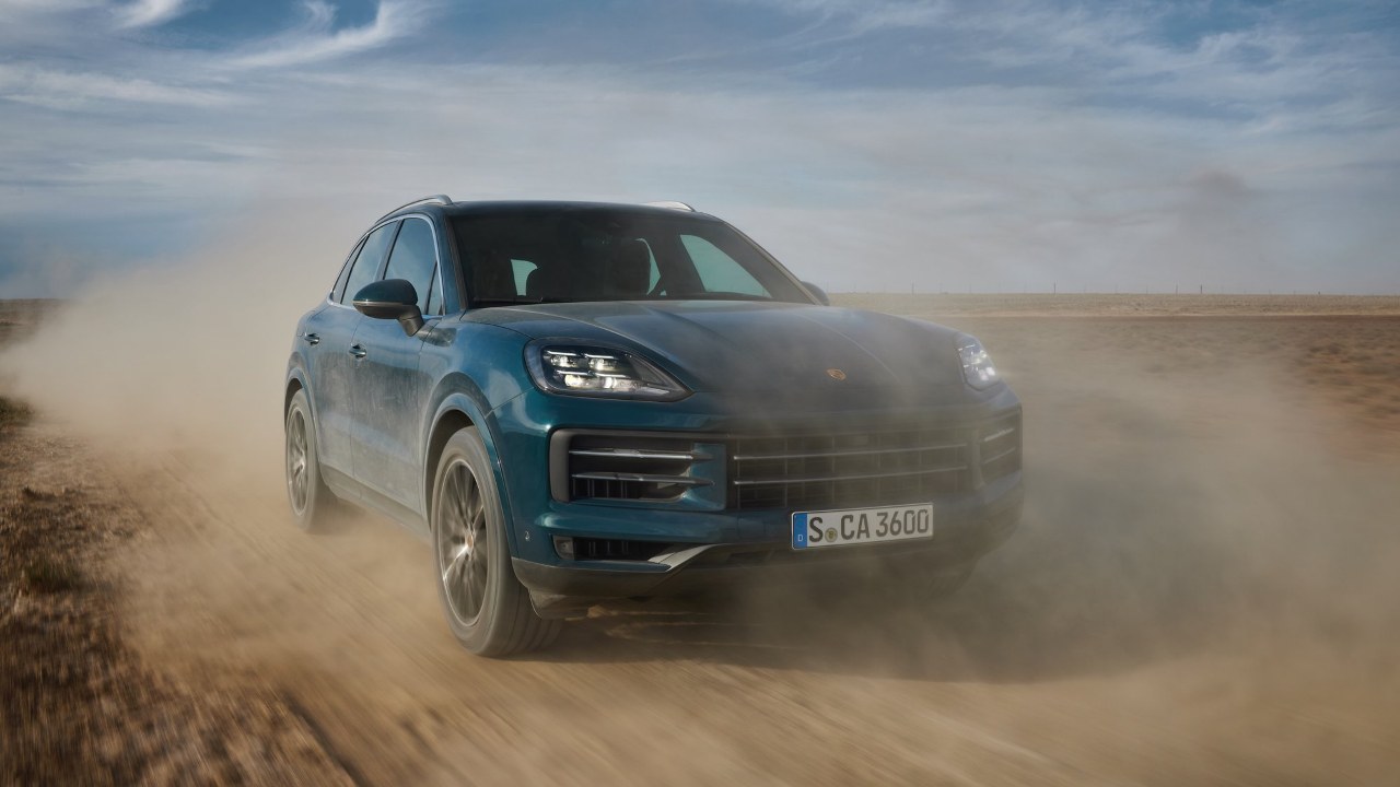 Porsche Cayenne SUV driving off‑road, kicking up dust from a front three‑quarter angle
