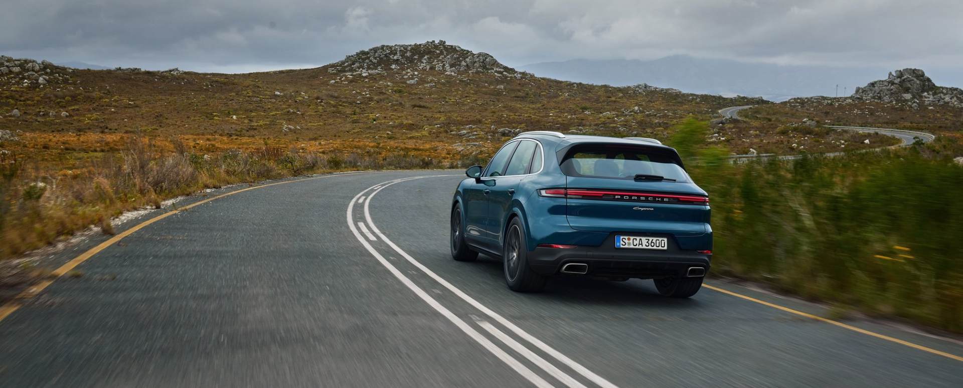Porsche Cayenne SUV driving away on a winding rural road, rear three‑quarter view