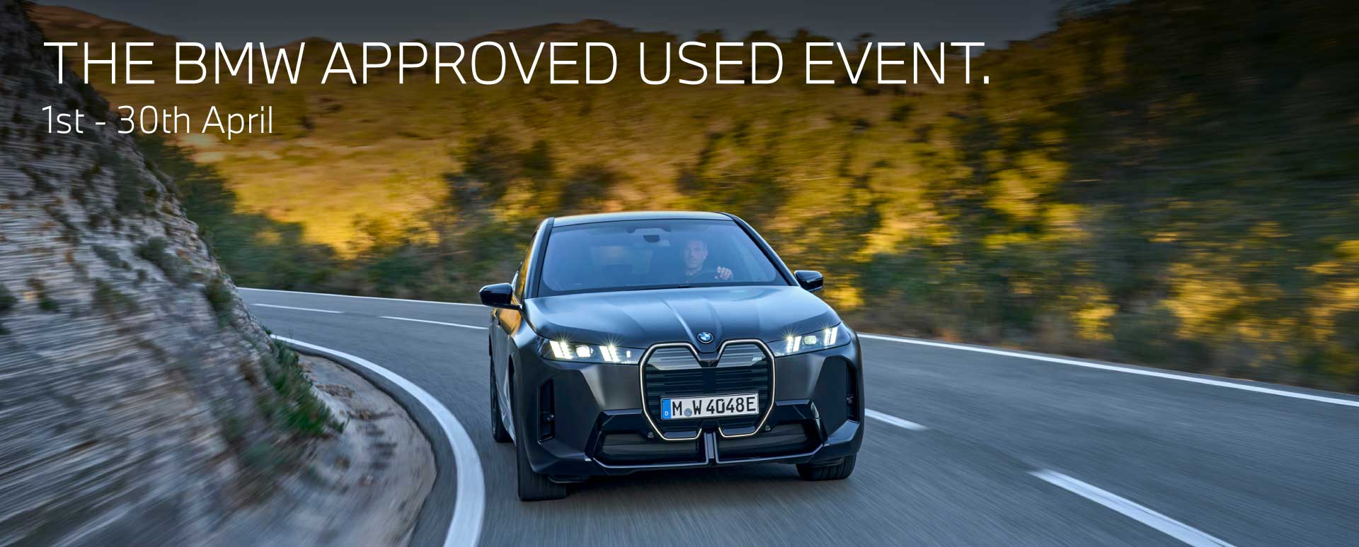 Front shot of black BMW vehicle driving on a road surrounded by trees. Promotional text overlay.