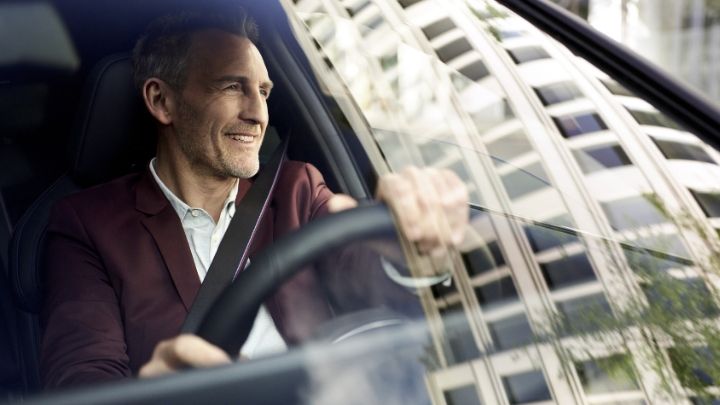 Man smiling whilst driving a BMW with reflection of a building of flats on the windscreen