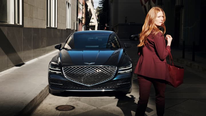 Blue Genesis G80 parked with woman walking past wearing a red suit