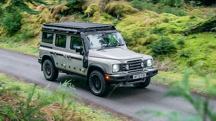A khaki green INEOS Grenadier driving down a road in a woodland