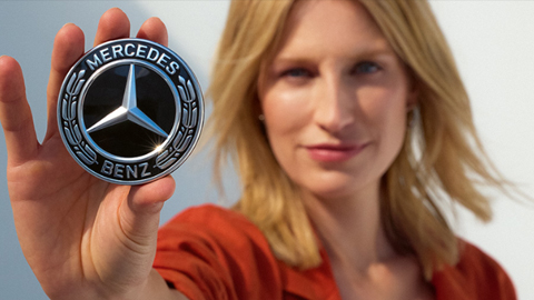 Blonde woman holding a Mercedes-Benz badge to the camera.